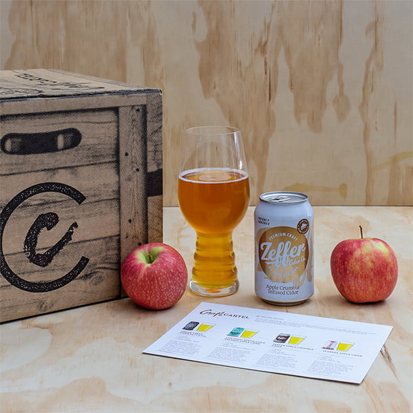 Zeller Apple Cider can, glass, and apples on a wooden surface with a crate in the background.