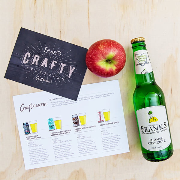 Green bottle of Frank's Summer Apple Cider with an apple and a 'Crafty Selection' card on a wooden surface.