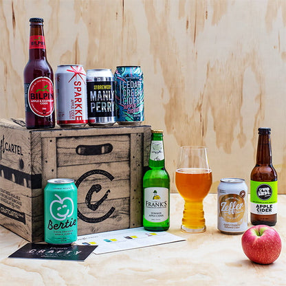 Assorted apple cider bottles and cans on a wooden crate with a glass of cider and an apple.